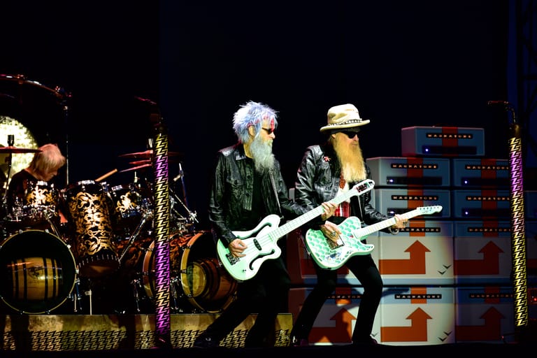 Two guitarists perform on stage under dramatic lighting with amplifiers and equipment visible behind them.