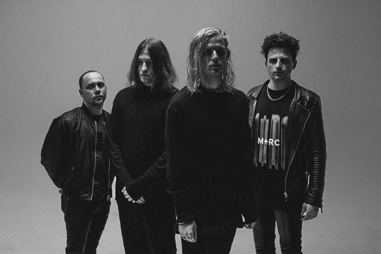Four band members stand against a grey background in a black and white promotional photograph.
