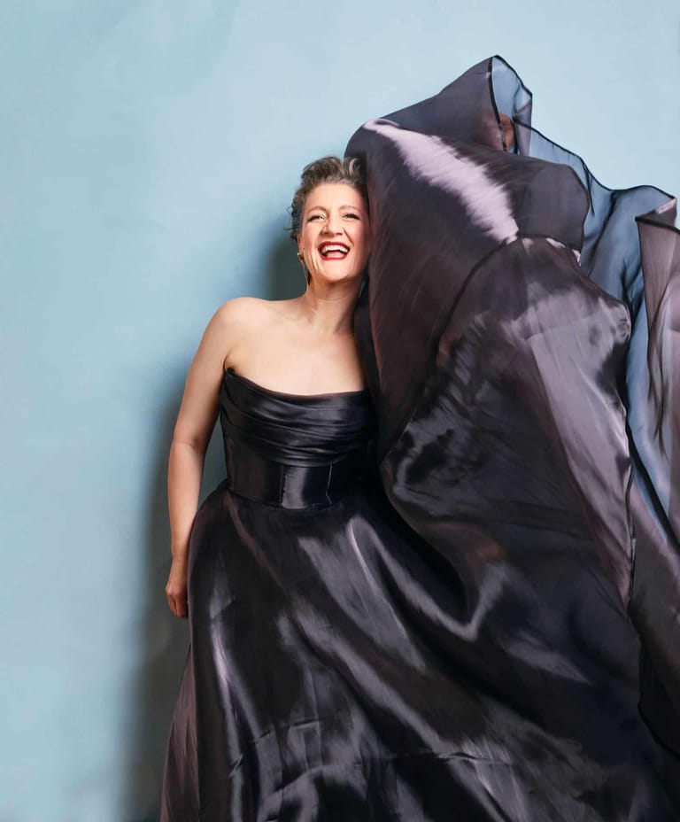 Woman in a black strapless gown with flowing fabric against a light blue background, smiling brightly.
