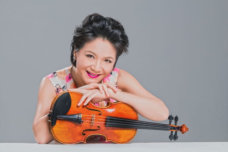Violinist with dark hair and red lipstick leaning on an amber violin against a grey background.