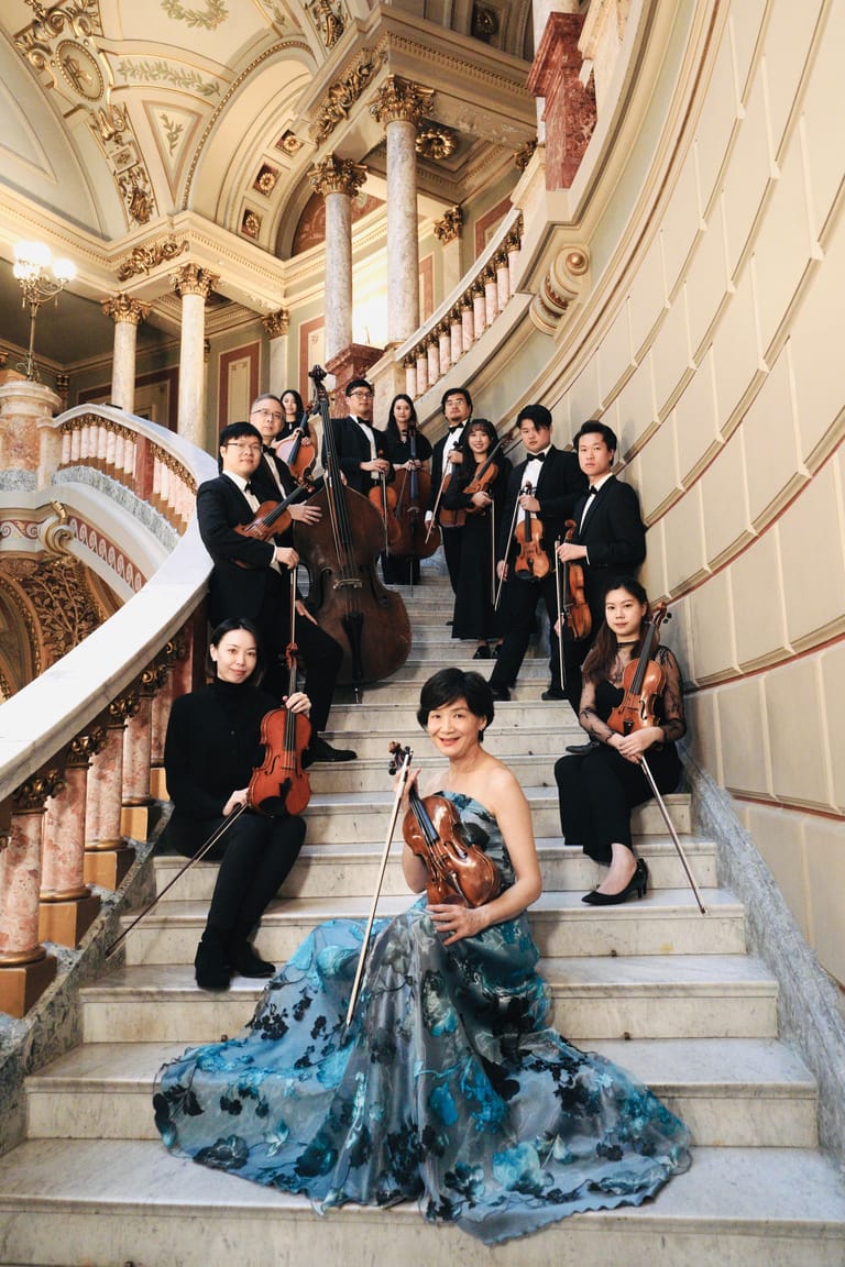 Orchestra members in formal black attire and a soloist in an elaborate blue gown pose on a grand ornate staircase.