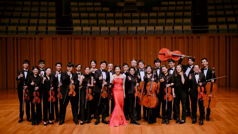Orchestra members in formal attire stand posed on a wooden stage with red curtains behind them.