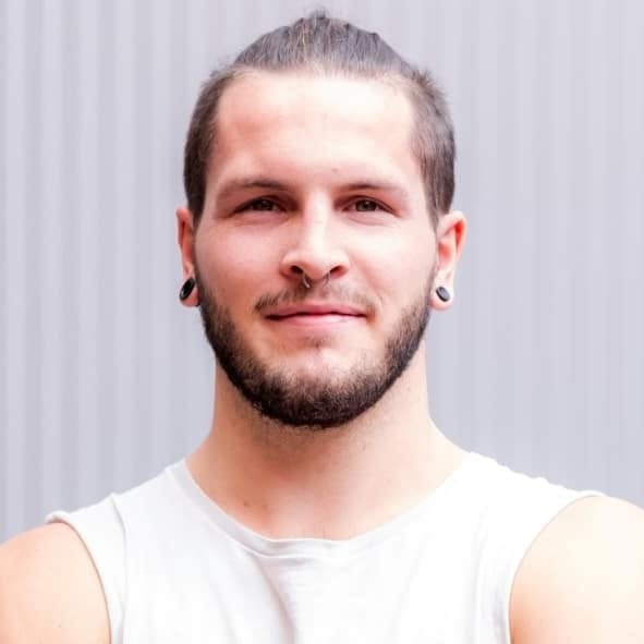 Man with dark hair and beard wearing a white sleeveless shirt against a white corrugated backdrop.