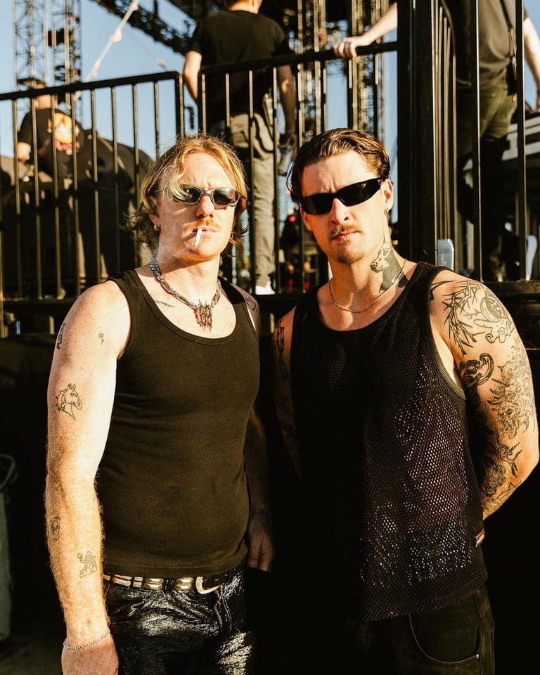 Two musicians in sunglasses and sleeveless black tops stand against an industrial metal backdrop in bright sunlight.