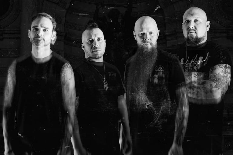 Four men in dark clothing stand against a black background in a monochrome promotional photograph.