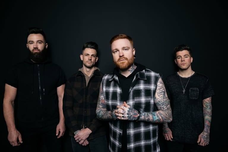 Four men stand against a dark background, the centre figure wearing a plaid shirt with visible arm tattoos.