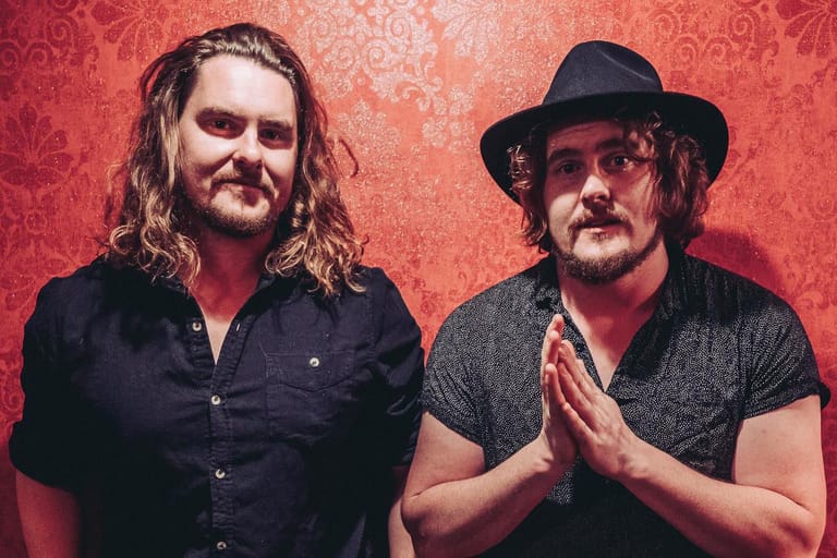 Two men with long hair and facial hair stand against a red damask patterned backdrop, one wearing a black shirt and the other a grey shirt with a black hat.