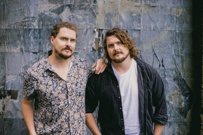 Two men with shoulder-length hair stand against a weathered grey wall in casual clothing.