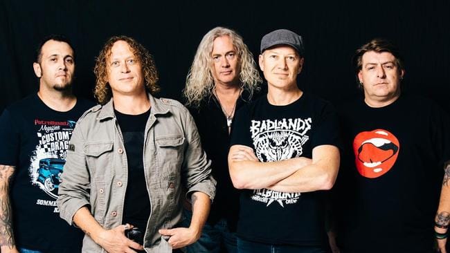 Five band members stand against a dark background, wearing casual black band t-shirts and varied styling.