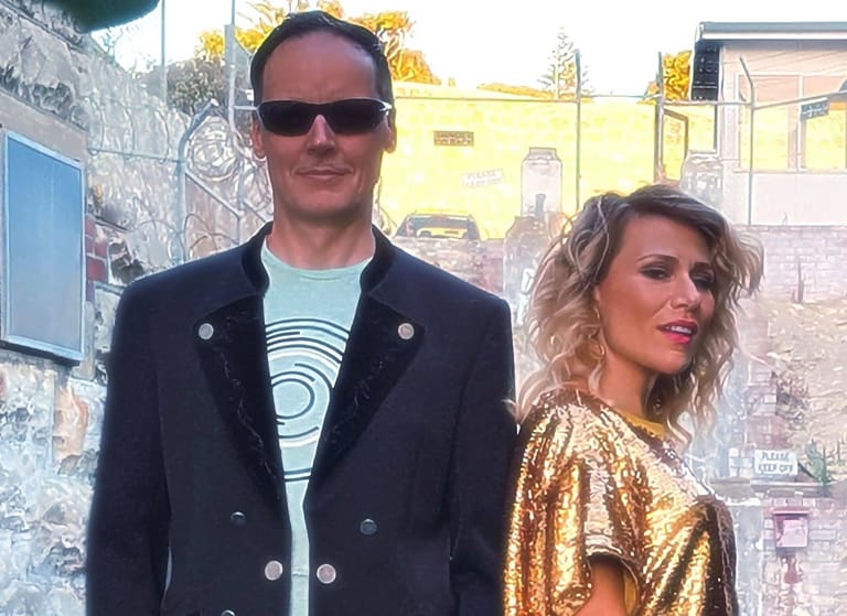 Two people stand outdoors against a weathered urban wall, one wearing a dark blazer and sunglasses, the other in a gold sequined top.