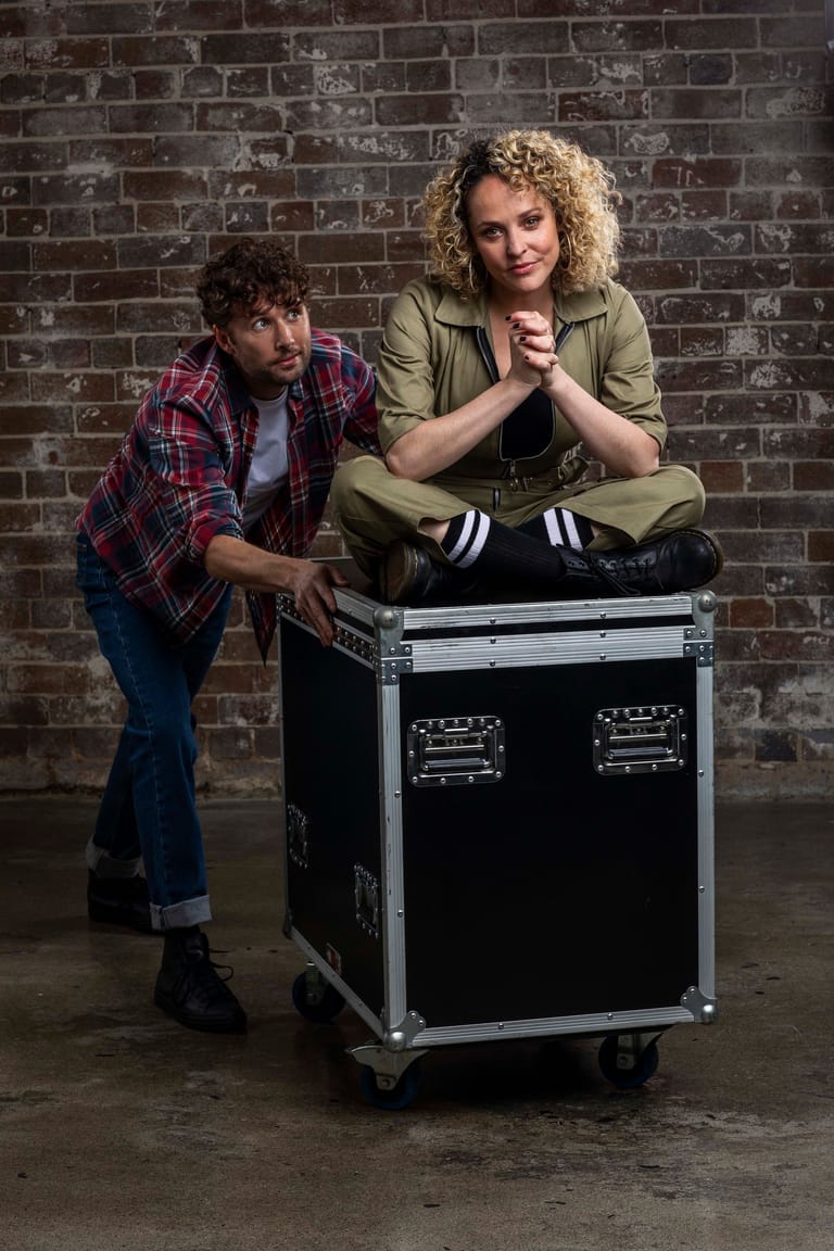 Two musicians pose against a brick wall beside a flight case, one seated atop it and one standing alongside.