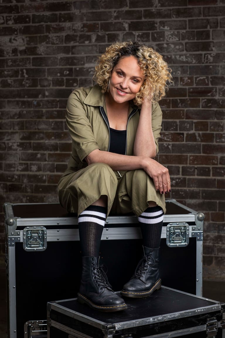 Person with curly blonde hair sitting on a flight case against a brick wall, wearing a khaki shirt and black boots.