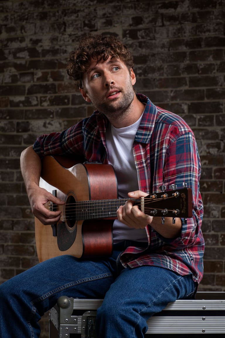 Musician in red and blue plaid shirt holding acoustic guitar against exposed brick wall.