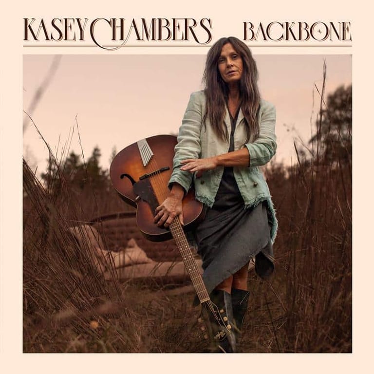 Musician holding a cello in a dry, overgrown field at dusk, wearing a cardigan and dark skirt.