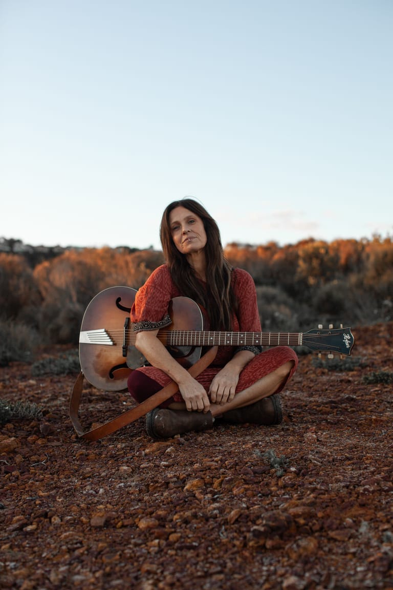 Woman in rust-coloured clothing seated cross-legged holding an acoustic guitar in an autumnal landscape.