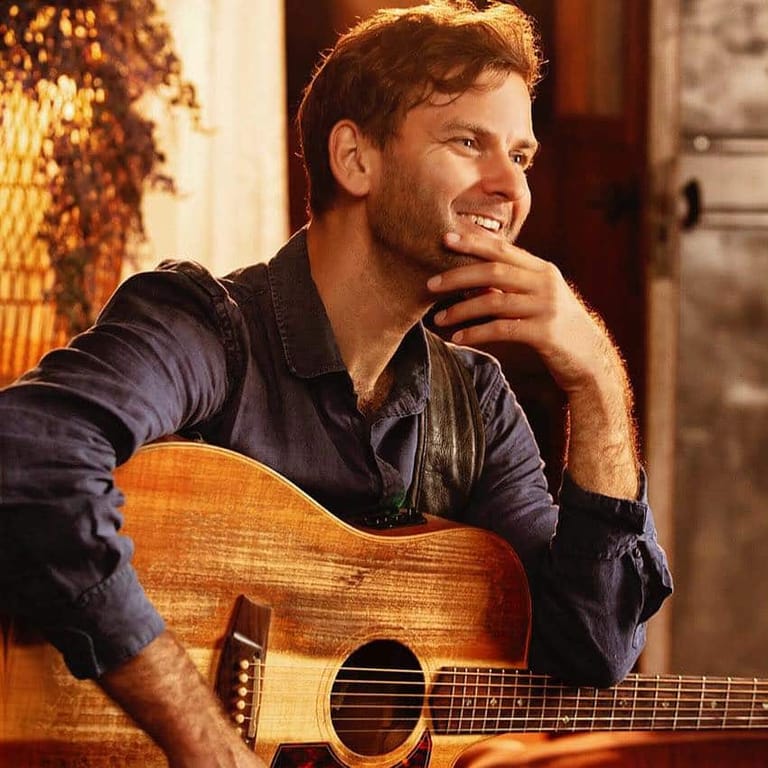 Musician smiling while holding an acoustic guitar in warm indoor lighting.