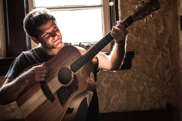 Musician playing acoustic guitar in natural window light, seated indoors.
