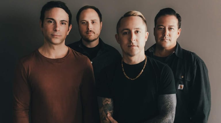 Four band members stand shoulder-to-shoulder against a neutral grey background in a promotional portrait.