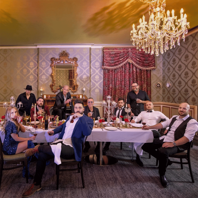 Ensemble cast gathered around a dining table in an ornate period room with crystal chandelier during a theatrical performance.