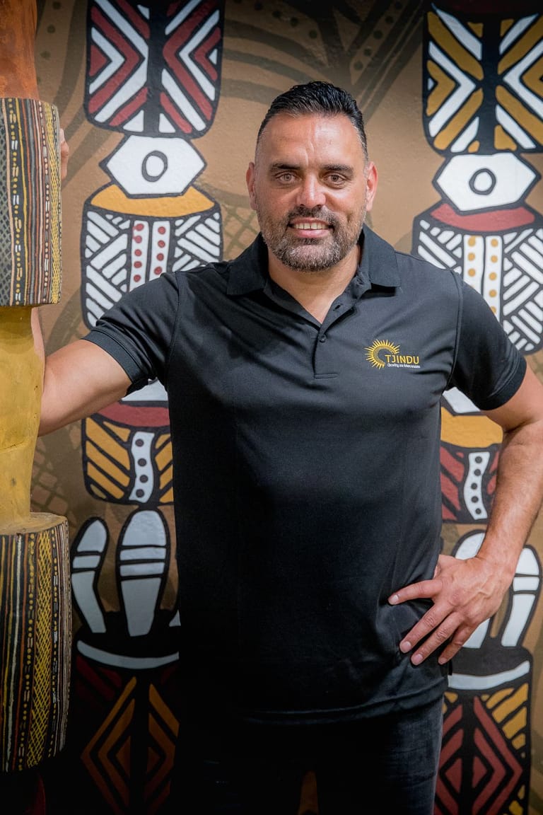 Man in black polo shirt posed against Aboriginal dot painting-style backdrop with geometric patterns in red, yellow and white.