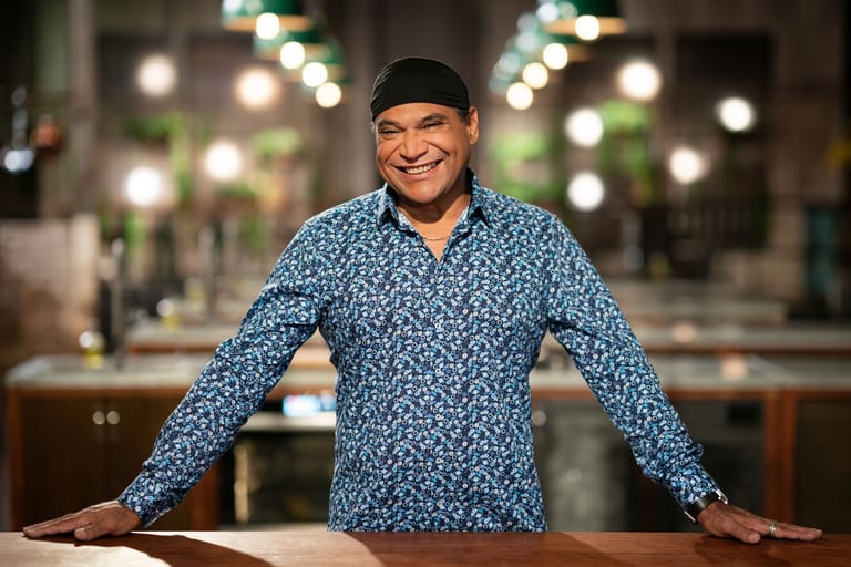 Man in blue patterned shirt and black beanie smiles at camera in front of blurred city lights.