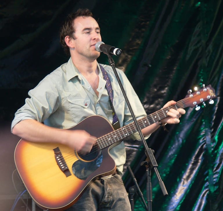 Musician singing into microphone while playing acoustic guitar on stage with green lighting backdrop.