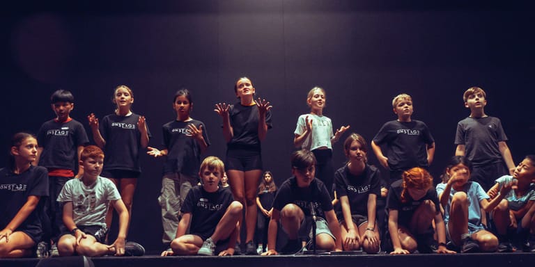 Large ensemble of performers in dark clothing arranged on stage under warm theatrical lighting.