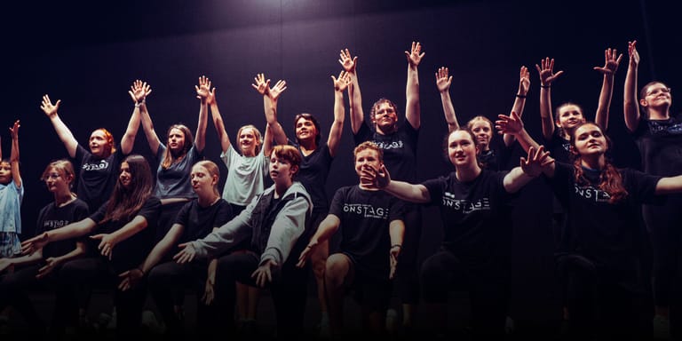 Large ensemble cast raising their arms in unified celebration on a darkly lit stage.