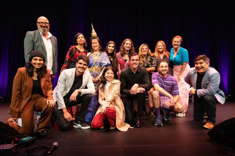 Large ensemble cast poses together on a stage lit with purple lighting during a theatrical production.