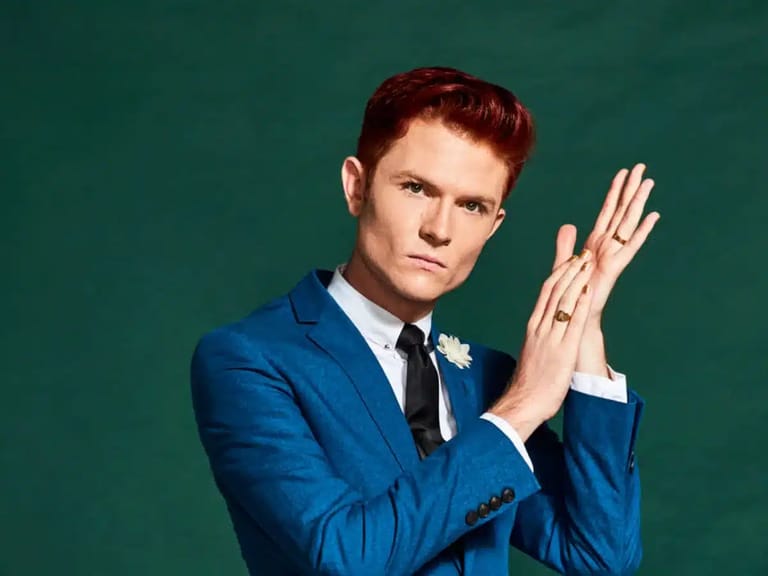 Man with reddish hair in a blue suit and tie gestures expressively against a teal-green background.
