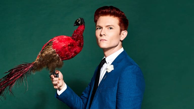 Man in a blue suit holding a red and gold macaw against a teal background.