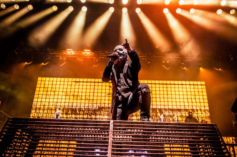 Performer in black clothing crouches on stage platform beneath dramatic golden spotlights and illuminated backdrop.