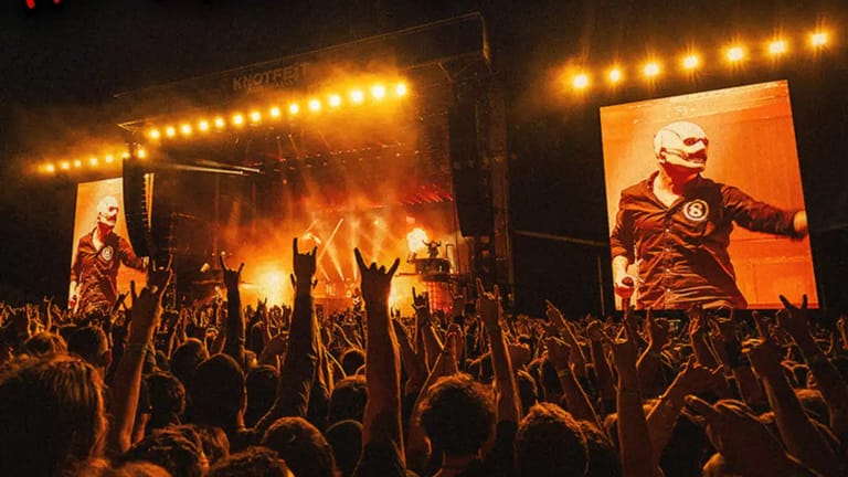 Large crowd with raised arms at an outdoor music festival stage lit by warm golden lights and giant screens.