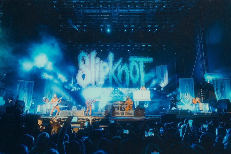 Band performing on stage beneath large illuminated logo with blue lighting and crowd in foreground.