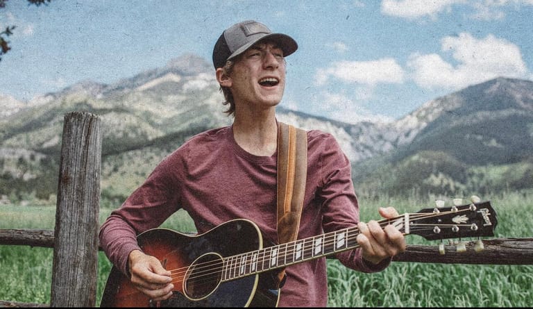 Musician with guitar smiling outdoors against mountain landscape and wooden fence.