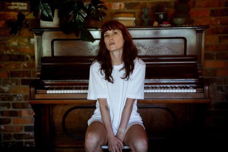 Woman with reddish-brown hair in white shirt seated at piano in brick-walled room.