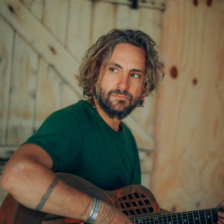 Man with shoulder-length hair and beard holding an acoustic guitar against a textured cream wall.