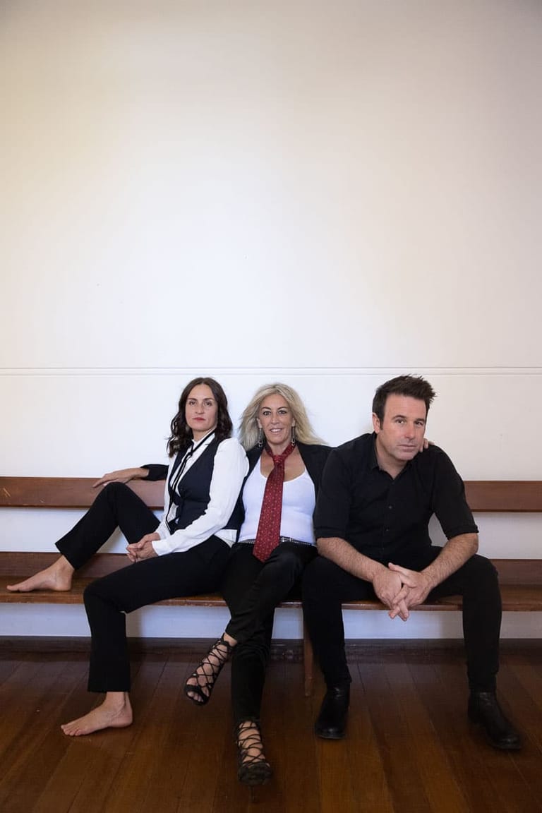Three performers in business attire sit casually on a wooden bench against a white wall.