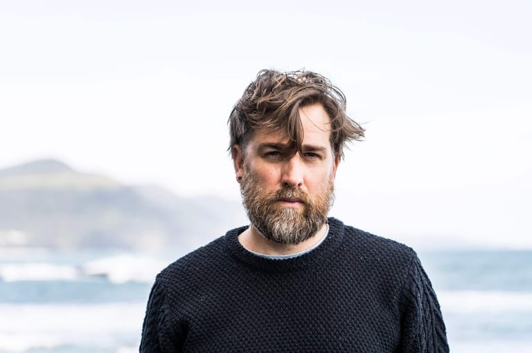 Man with beard in dark sweater standing before coastal seascape with overcast sky.