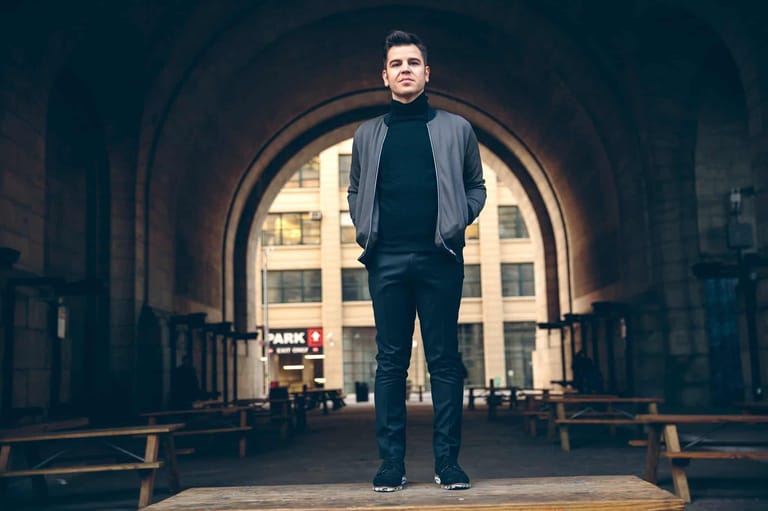 Man standing in archway of brick corridor, backlit by warm city light.