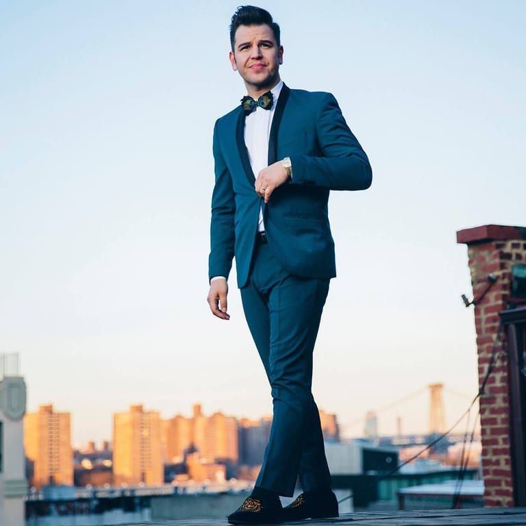 Man in teal suit and bow tie standing against urban skyline at sunset.