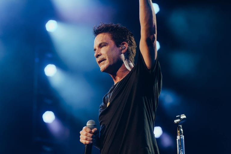 Performer on stage holding a microphone with arm raised, illuminated by blue stage lighting.