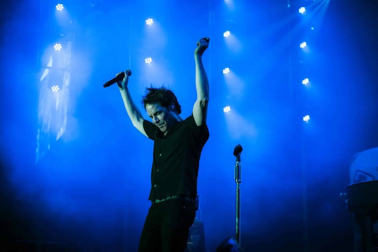 Performer on stage with arms raised, holding microphones, under blue stage lighting with scattered white lights.