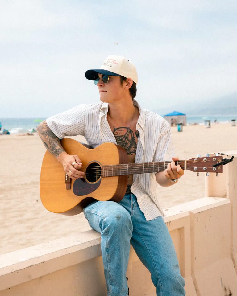 Musician with acoustic guitar sitting on a beach bench, wearing a cream cap and striped shirt, ocean and sand visible behind.