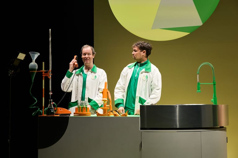 Two performers in white coats and green scarves stand at a laboratory-style desk during a stage production.