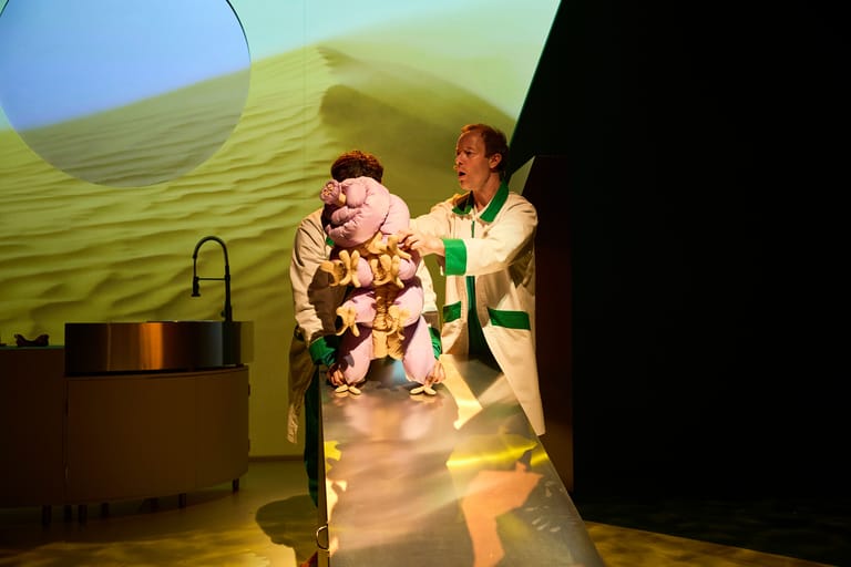 Two performers on stage with a child, illuminated by yellow-green lighting beneath a projected landscape backdrop.