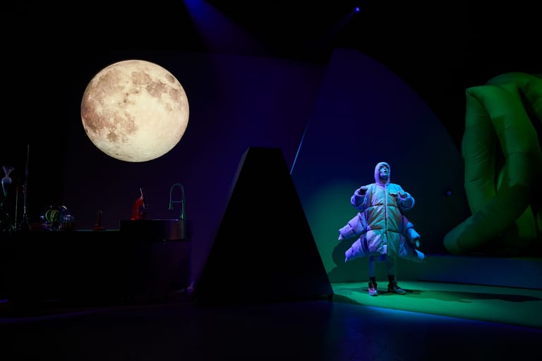 Performer in blue lighting on stage beneath a large illuminated moon against dark backdrop.