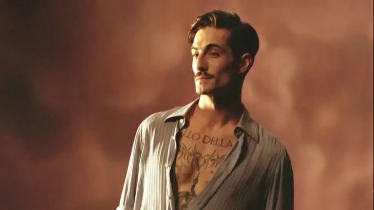 Man with tattooed chest in open grey shirt against warm-toned atmospheric backdrop.