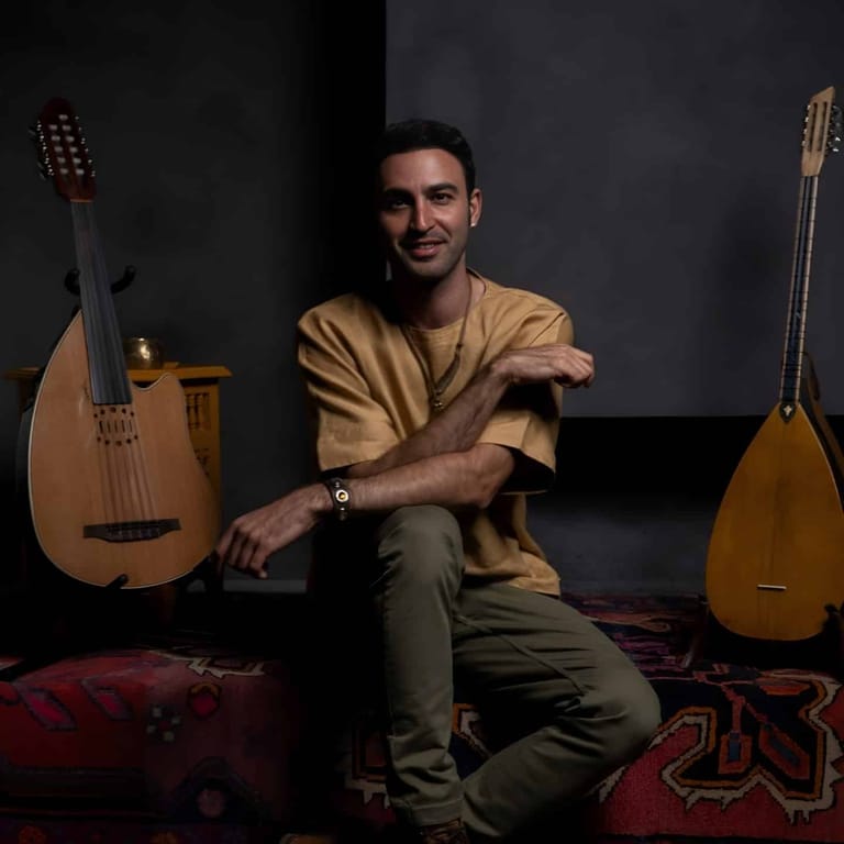 Musician seated between two classical guitars against a dark backdrop, wearing a tan shirt and smiling at the camera.