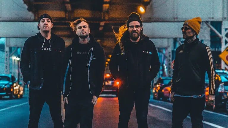 Four band members stand on an urban street at dusk, lit by warm streetlights and vehicle headlights behind them.